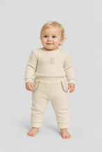 Load image into Gallery viewer, A baby standing and wearing a cream-colored ribbed knit two-piece outfit consisting of a long-sleeve top and matching pants, photographed against a solid light gray background.